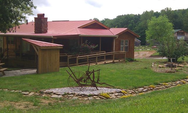 Star Falls Resort Cabin on a cloudy spring day. The front of the cabin is shown. The front yard has bright green grass with a small structure and an antique farming implement.
