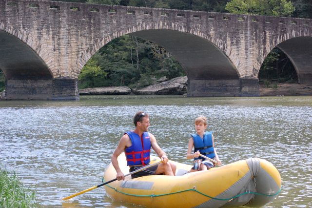 A father and son rafting on a beautiful summer day. The river is calm and a bridge can be seen behind them. 